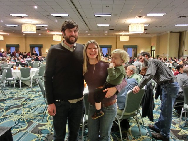 A family of three posing at an indoor event with many seated attendees.