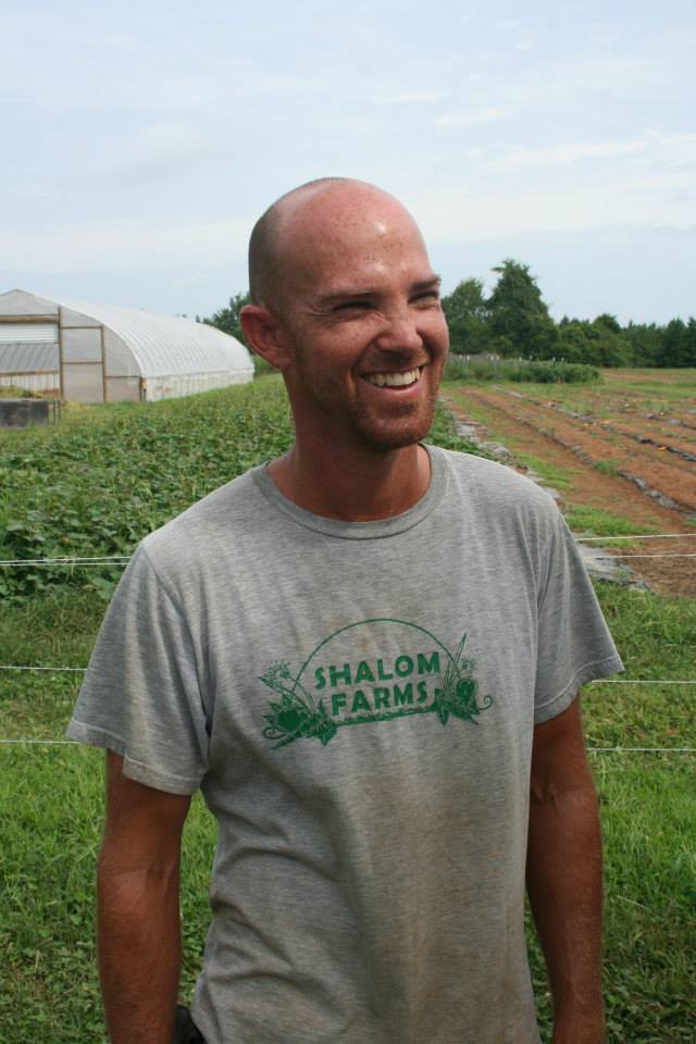 A man smiling in a farm field wearing a Shalom Farms t-shirt.