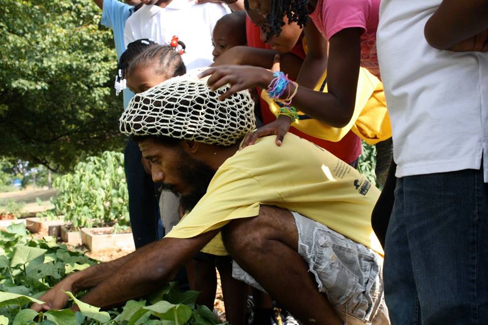 A boy crouching outdoors, wearing a yellow shirt and a patterned hat.