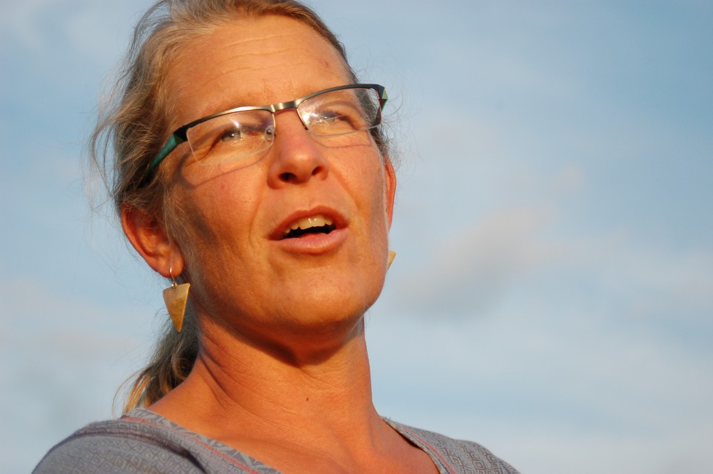 A woman with glasses speaking outdoors against a blue sky.