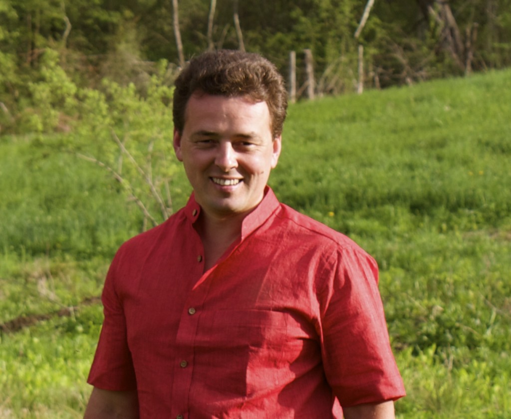 A man in a red shirt standing outdoors with greenery.