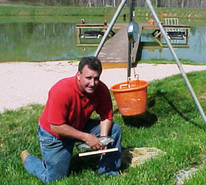 Man in red shirt kneeling by a campfire setup near water.