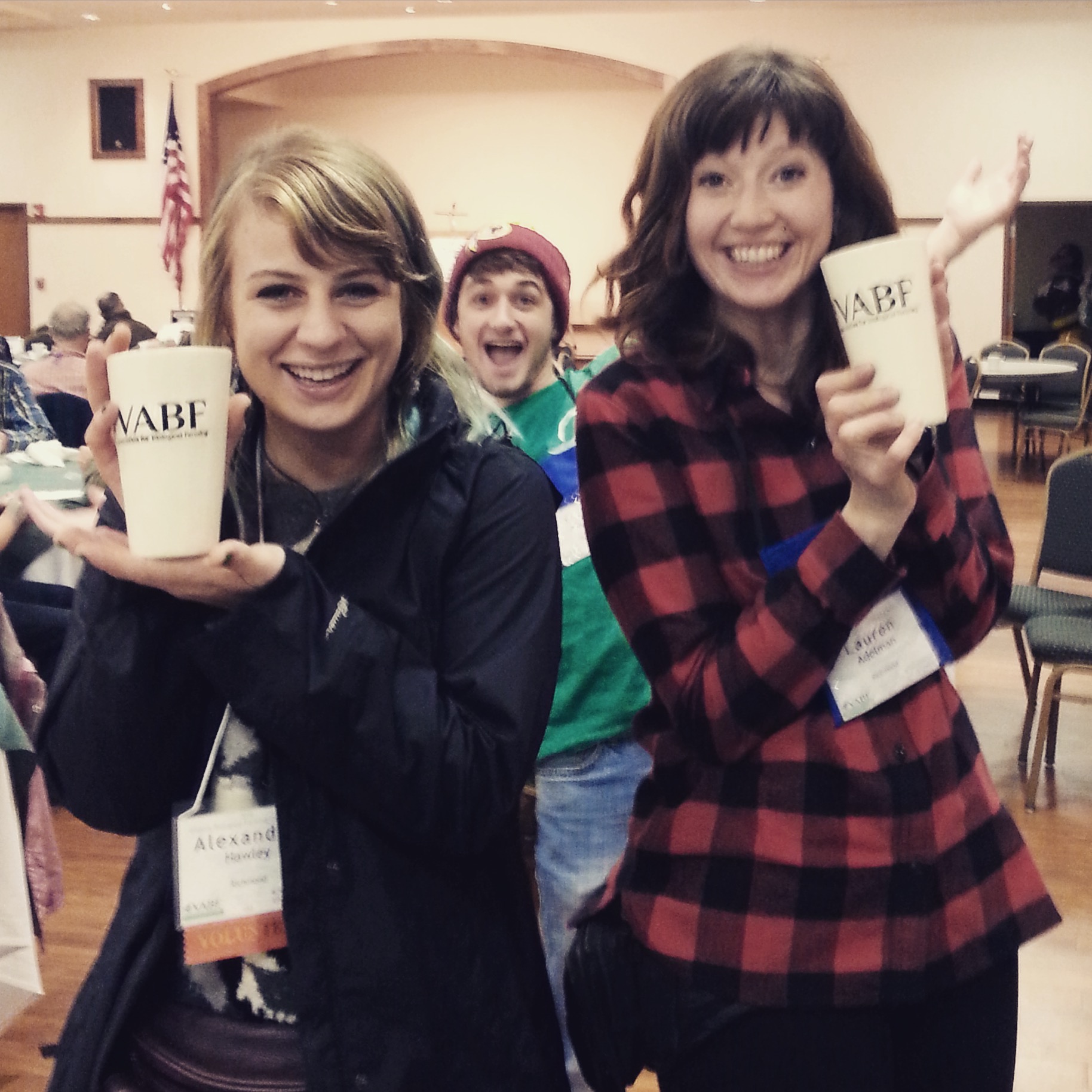 Two women smiling and holding cups while a man photobombs in the background.