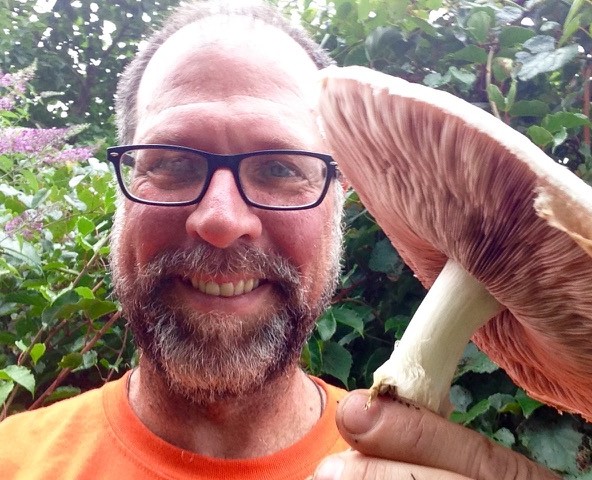 A man smiling while holding a large mushroom outdoors.