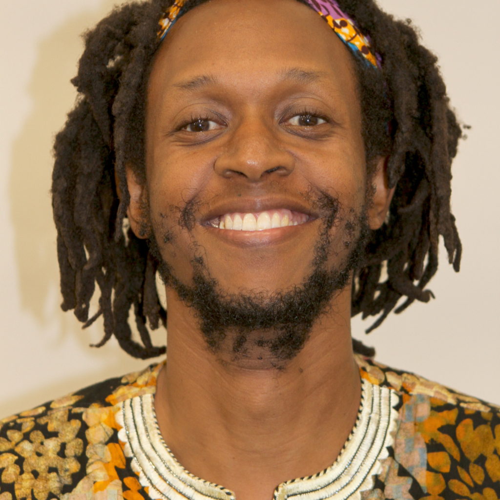 A smiling man with dreadlocks wearing a patterned shirt.