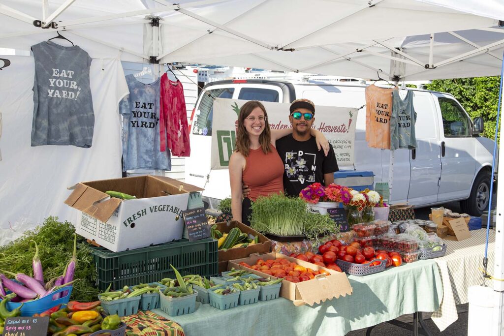 Two vendors at a fresh produce market stall under a white canopy.