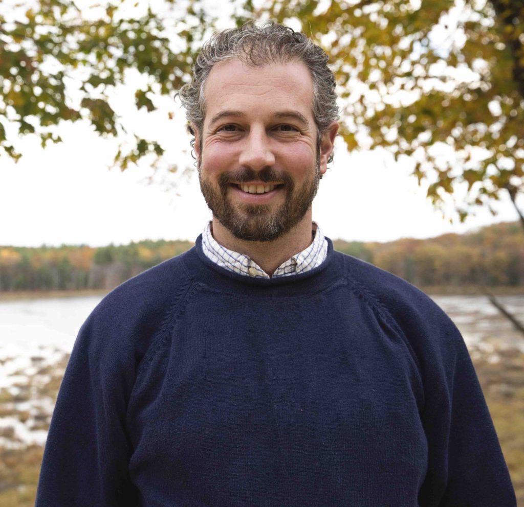 A smiling man outdoors in a navy sweater with autumn trees behind.