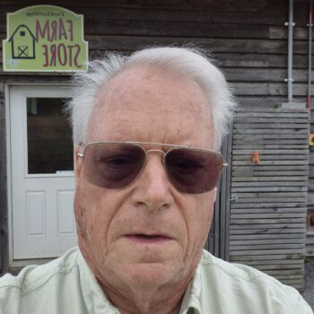 An elderly man wearing sunglasses poses outside a rustic building.