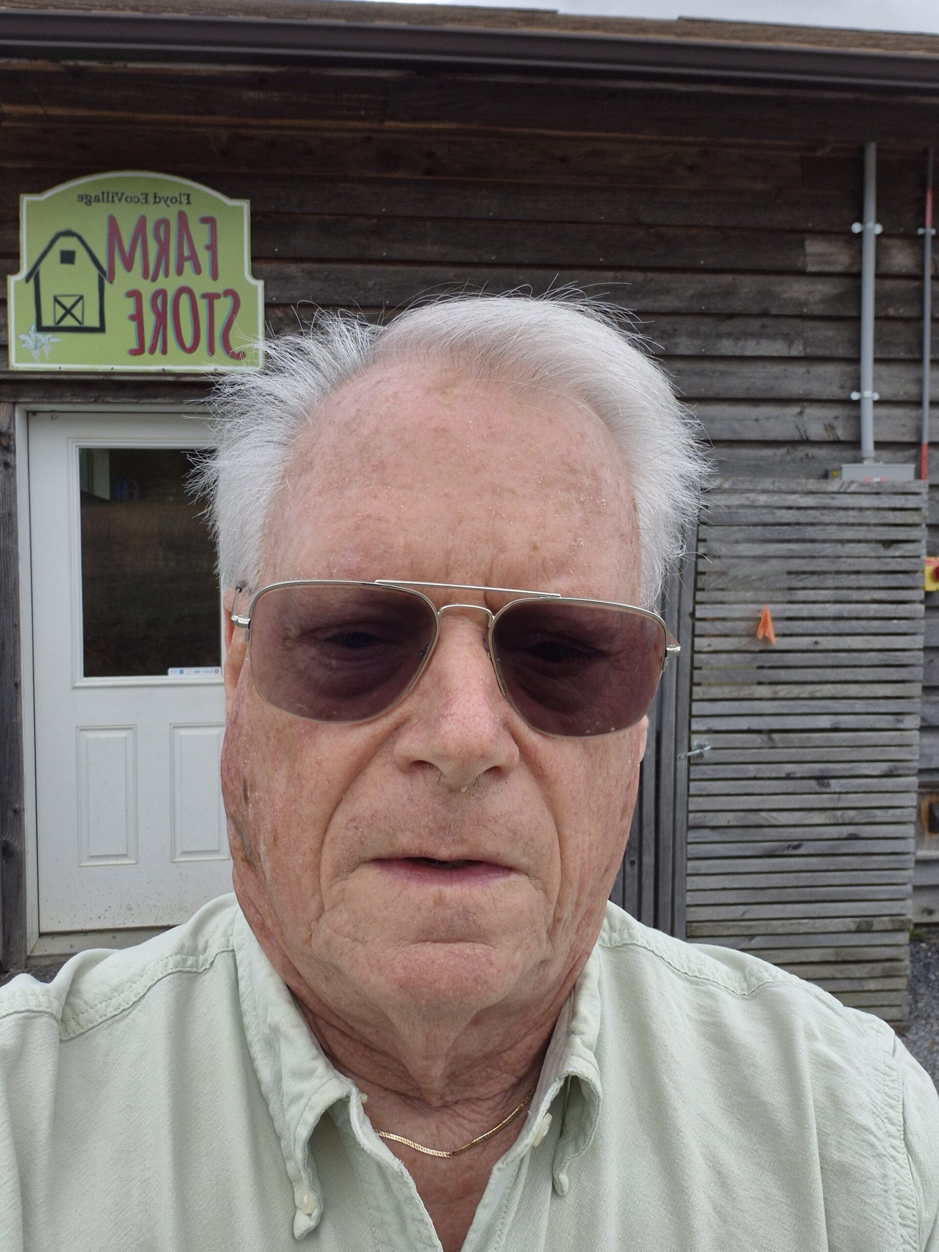 An elderly man wearing sunglasses poses outside a rustic building.
