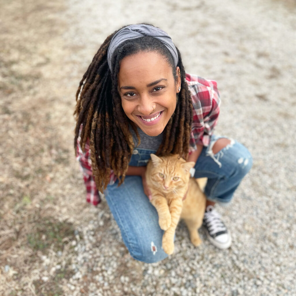 A smiling woman crouches outdoors holding an orange cat.