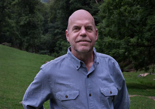 A man in a blue shirt stands outdoors with greenery behind him.