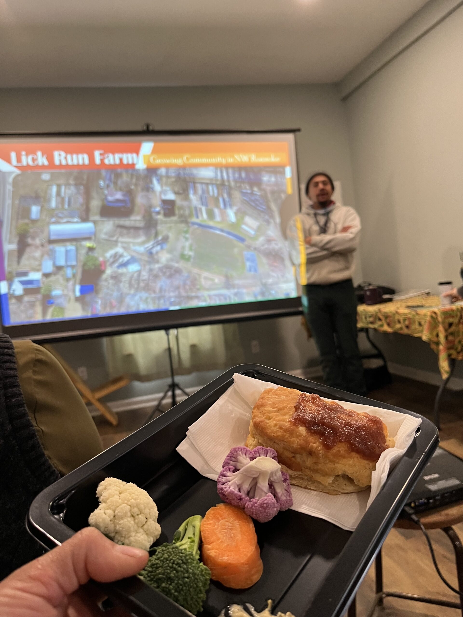 A man stands in front of a screen showing a live feed of a market, with food in the foreground.