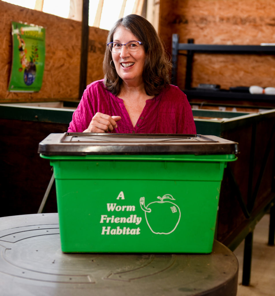 A woman smiling behind a green recycling bin.